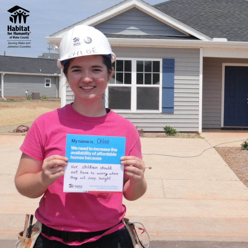 Woman in white construction helmet and pink shirt holds a sign that says "We need to increase the availability of affordable homes because our children should not have to worry where they will sleep tonight. 