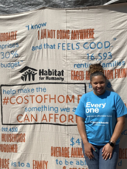 Deanna is wearing a Blue t-shirt that says Everyone deserves an affordable home in front of a canvas that says help make the cost of home something we can all afford.